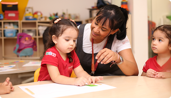 Professora ajudando uma aluna a desenhar enquanto outra criança observa